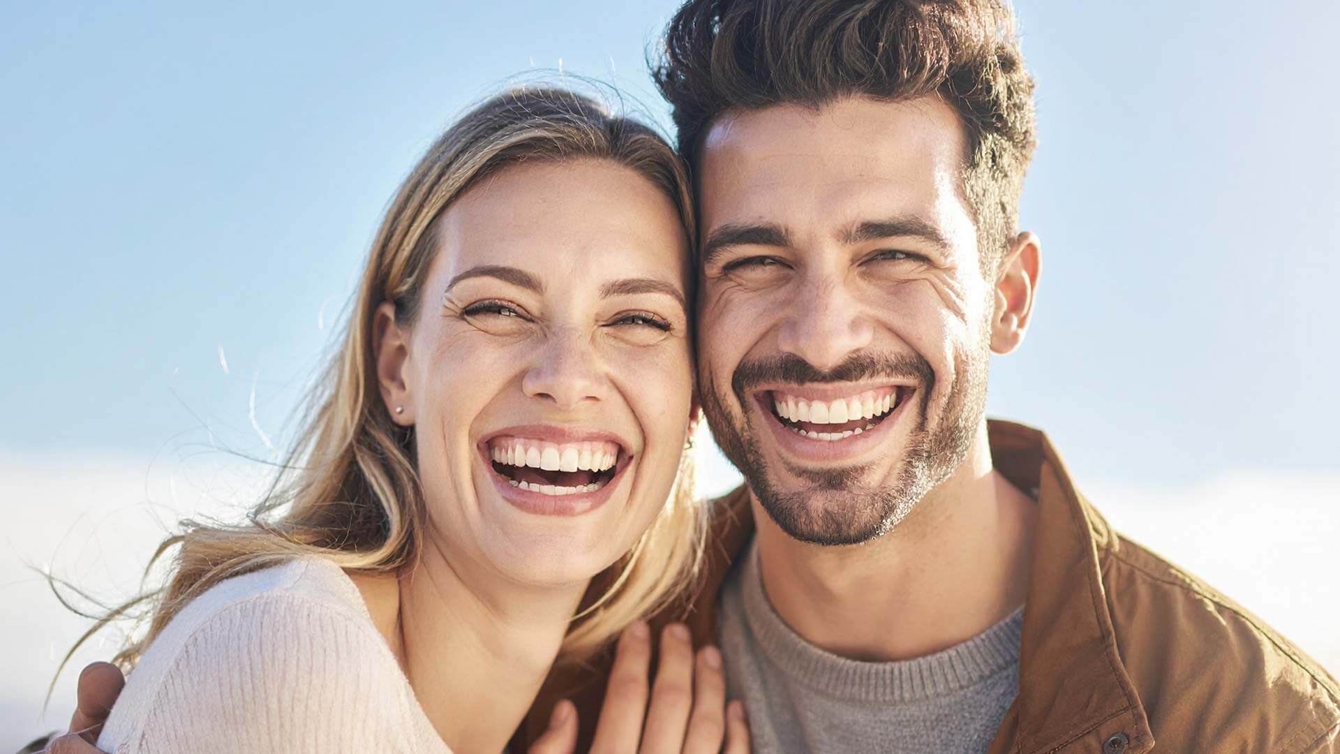 Junges Paar lächelt vor blauem Himmel und Sonnenschein in die Kamera